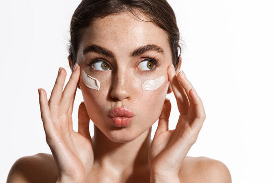 A playful shot of a young woman applying cream under her eyes, showcasing smooth and hydrated skin, highlighting DermaSkin’s Struck Gold Under-Eye Patches